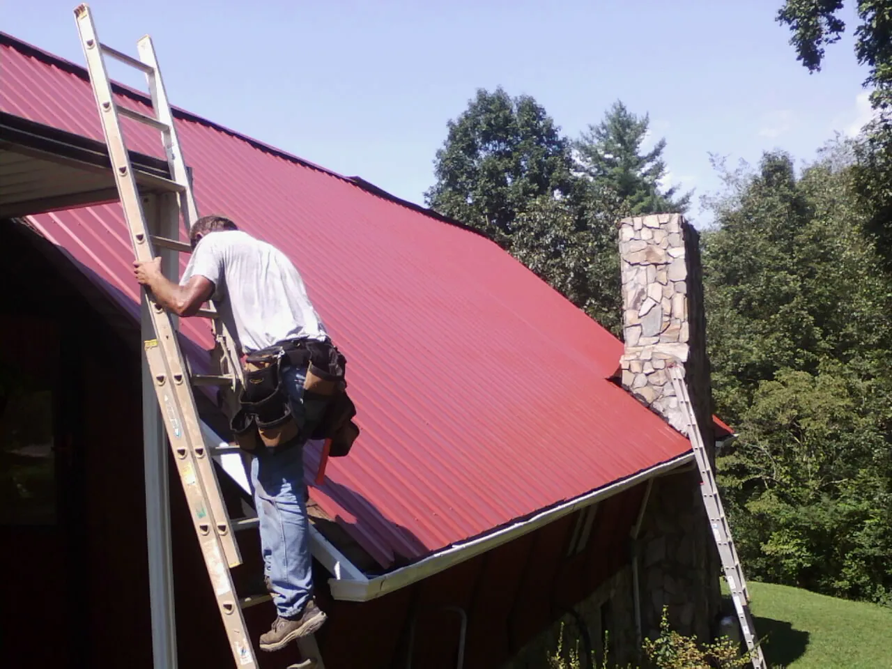 Roofer on a ladder beside a red metal roof and stone chimney — Soffit & Fascia Repair by Roofing Brothers in Calumet City